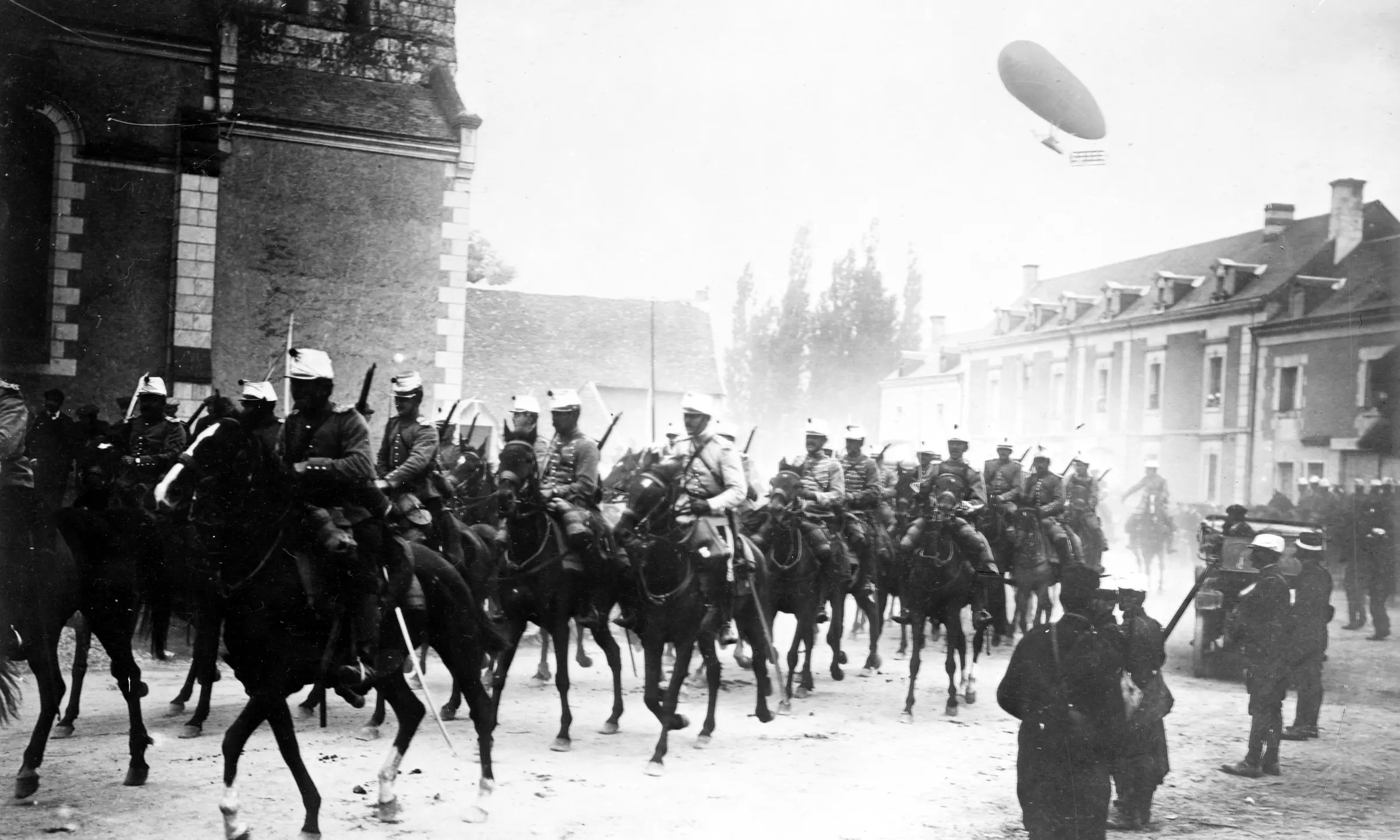 French cavalry ride through a town street while the army dirigible Dupuy de Lome flies overhead, 1914
